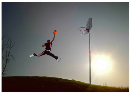 A basketball player is captured midair against a clear sky, legs spread wide in a split, holding an orange basketball high above his head as he approaches the hoop. The sun glows brightly to the right, casting dramatic light across the scene.