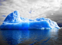 Left, a large, bright blue iceberg floats in calm, reflective water under a cloudy gray sky. The ice has jagged ridges and varying shades of blue, with a more intense hue near the waterline. Right, the same iceberg as in the previous image, but with altered color saturation. The blues in the ice and water are much more intense, while the sky and clouds have been desaturated to near black-and-white, creating a striking contrast.
