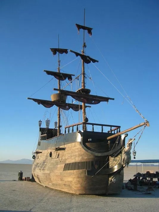 A large wooden ship styled like an old sailing vessel with three masts and black sails sits on dry land under a clear blue sky. The ship has a carved figurehead at the bow and appears stranded on a flat, desert-like surface, with small objects and people gathered nearby.