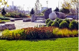 A landscaped park scene with green grass, flowering plants, and neatly trimmed bushes in the foreground. In the background, a stone sculpture installation features large geometric shapes, including a prominent spherical stone resting within curved stone forms. Trees and industrial buildings are visible in the distance.