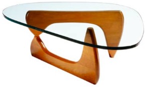 A modernist coffee table with a clear, irregularly shaped glass top. The base is sculptural, made of smooth, curved wood in a warm brown tone, forming two interlocking supports that balance the glass surface. The design is sleek and organic, combining functionality with artistic form.